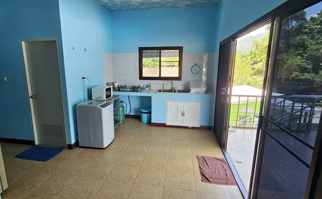 A view of an apartments open kitchen layout with tinted french windows opening to a first floor balcony. In the kitchen is a washing machine, microwave, gas hob, sink. There is a corridor opening to the left leading to a bathroom and bedrooms.