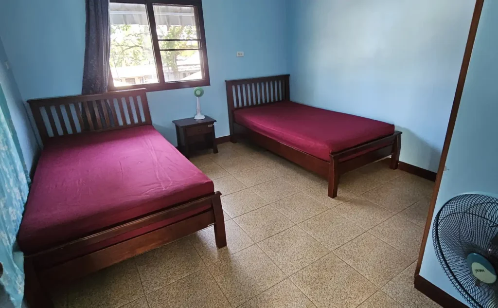 View of a double bedroom with two wooden beds, a bedside table with lamp, a floor fan for cooling and a window with view to a tree