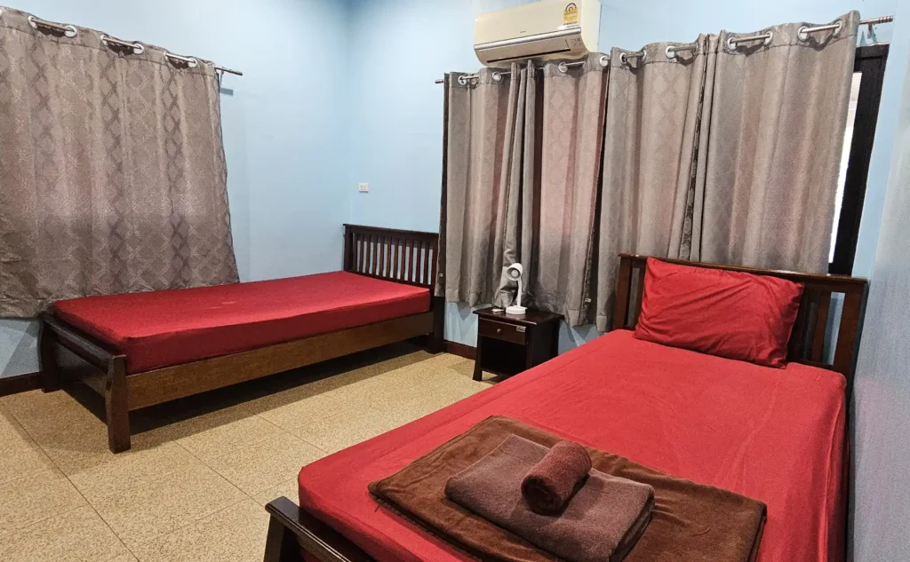 View of a double bedroom with two wooden beds, a bedside table with lamp. An air conditioning unit and two curtained windows giving a double aspect to the room. The curtains are closed